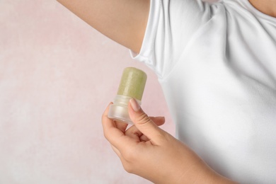 Young woman holding natural crystal alum deodorant near armpit on light pink background, closeup