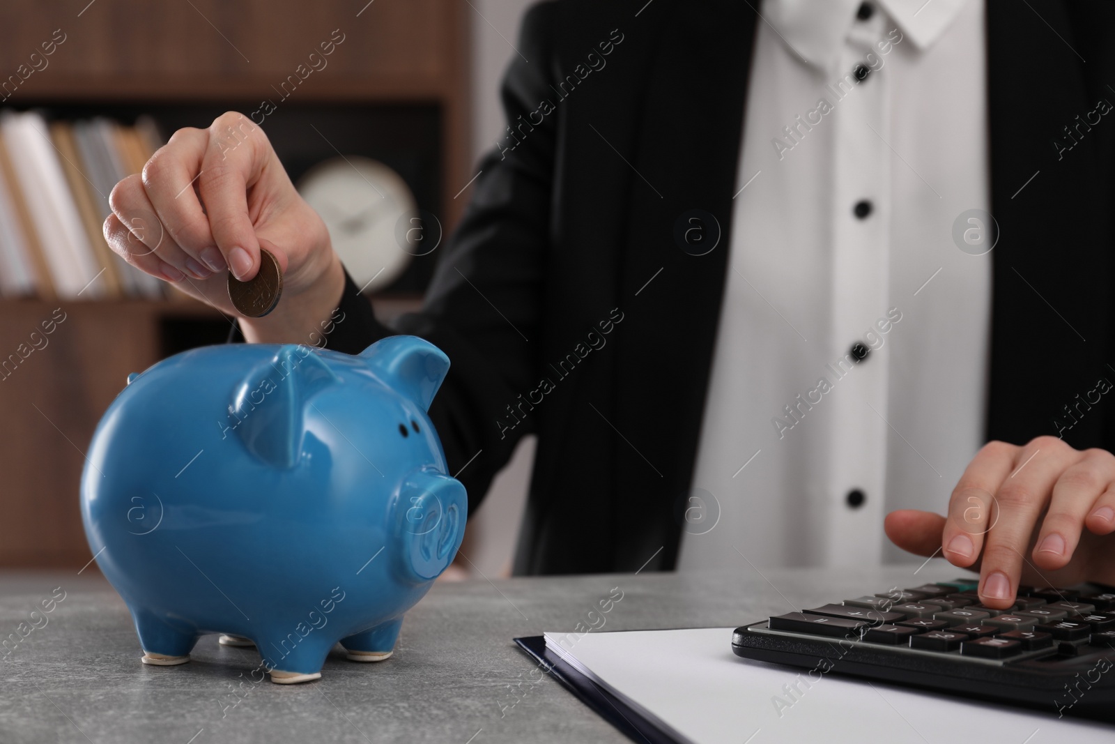 Photo of Woman with calculator putting coin into piggy bank at grey table indoors, closeup