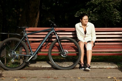 Photo of Woman with injured knee on wooden bench near bicycle outdoors