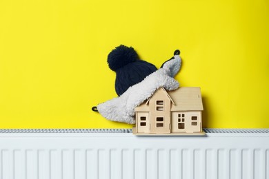 Photo of Wooden house model and hat on heating radiator near yellow wall. Energy efficiency concept