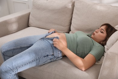 Young woman struggling to squeeze into tight jeans while lying on sofa at home