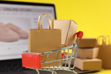 Photo of Online store. Mini shopping cart, purchases and laptop against yellow background, selective focus