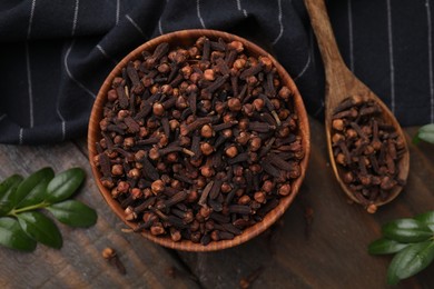 Photo of Aromatic cloves in bowl, spoon and green leaves on wooden table, flat lay