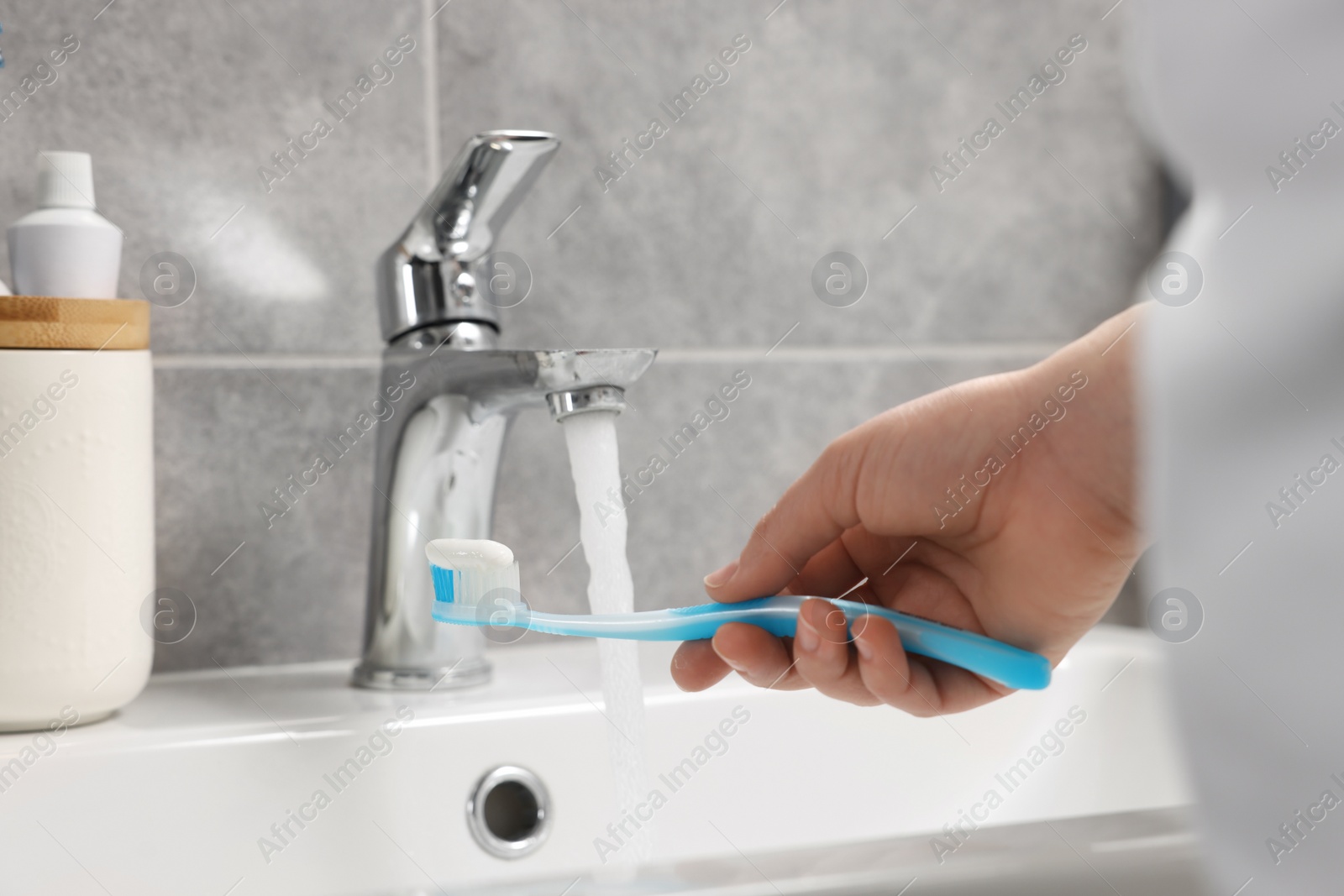 Photo of Woman holding plastic toothbrush with paste near flowing water above sink in bathroom, closeup