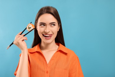 Happy young woman holding sushi roll with chopsticks on light blue background. Space for text