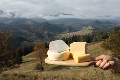 Woman holding board with different types of delicious cheeses against mountain landscape, closeup