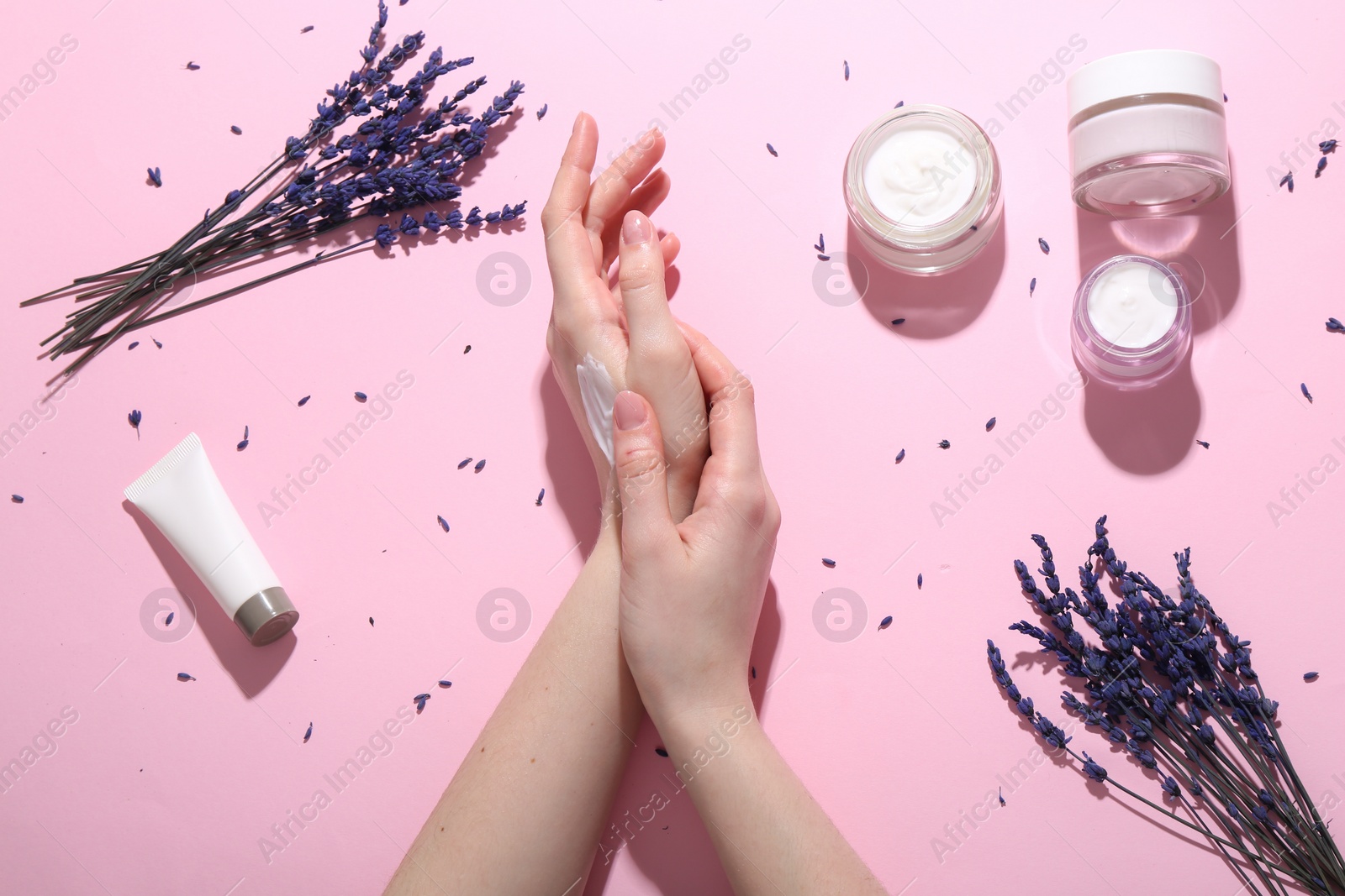 Photo of Woman applying hand cream and lavender flowers on pink background, top view