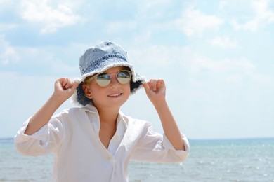 Little girl wearing sunglasses and hat at beach on sunny day. Space for text