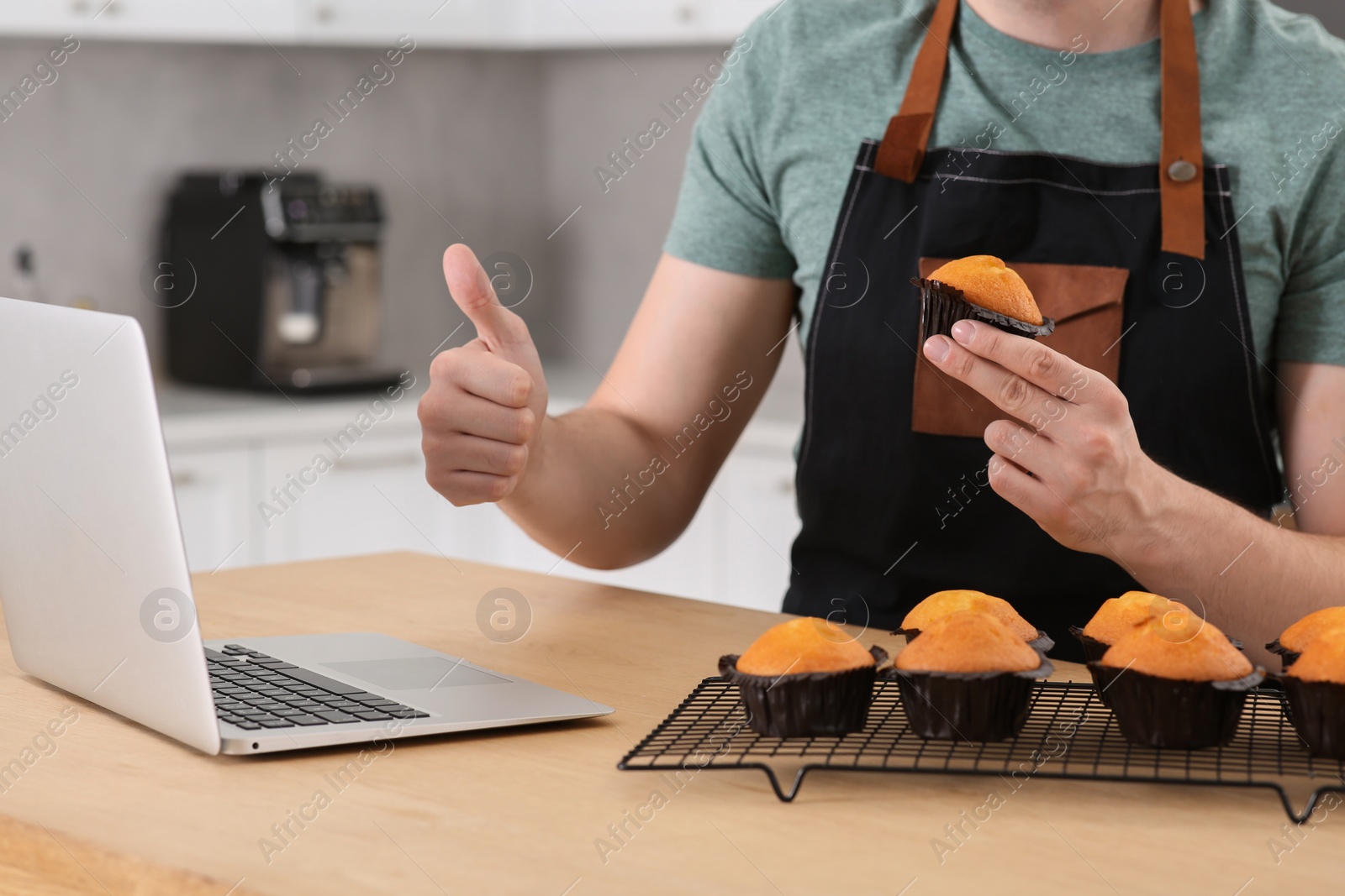 Photo of Man holding muffin near laptop and showing thumb up at table in kitchen, closeup. Time for hobby