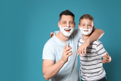 Photo of Happy dad and son with shaving foam on faces against blue background, space for text