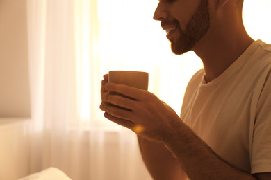 Photo of Young man with cup of coffee near window at home, closeup. Lazy morning