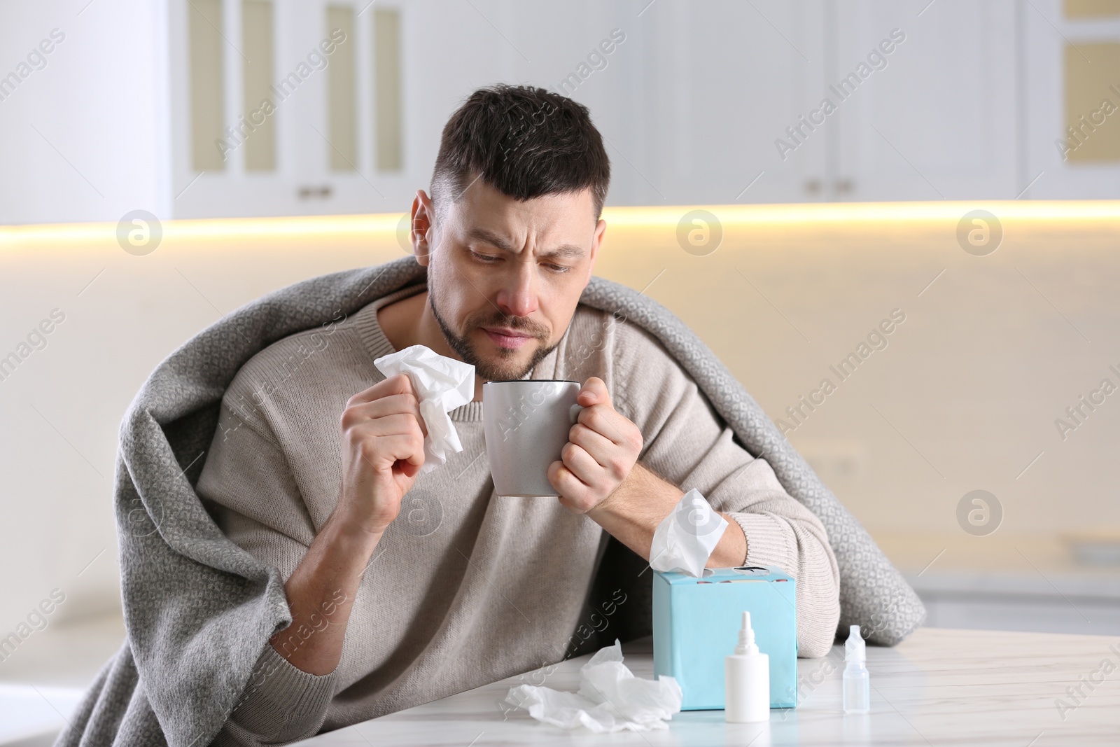 Photo of Ill man at table with nasal spray, drops and box of paper tissues in kitchen