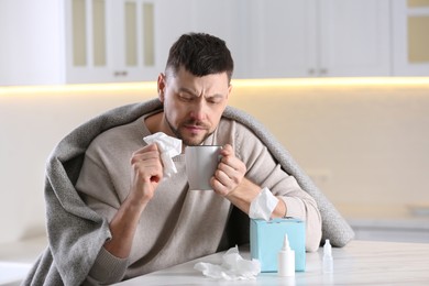 Photo of Ill man at table with nasal spray, drops and box of paper tissues in kitchen