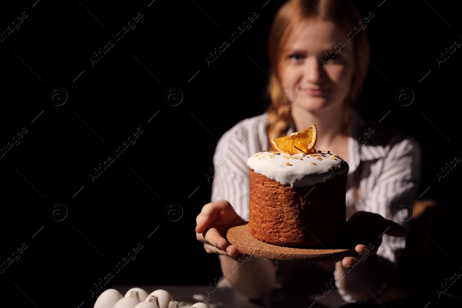 Photo of Young woman holding traditional decorated Easter cake against black background, focus on hands. Space for text