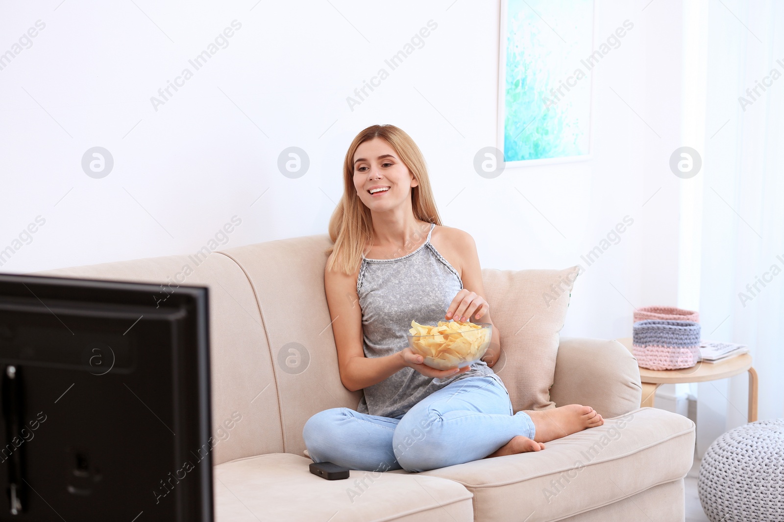 Photo of Woman with bowl of potato chips watching TV on sofa in living room