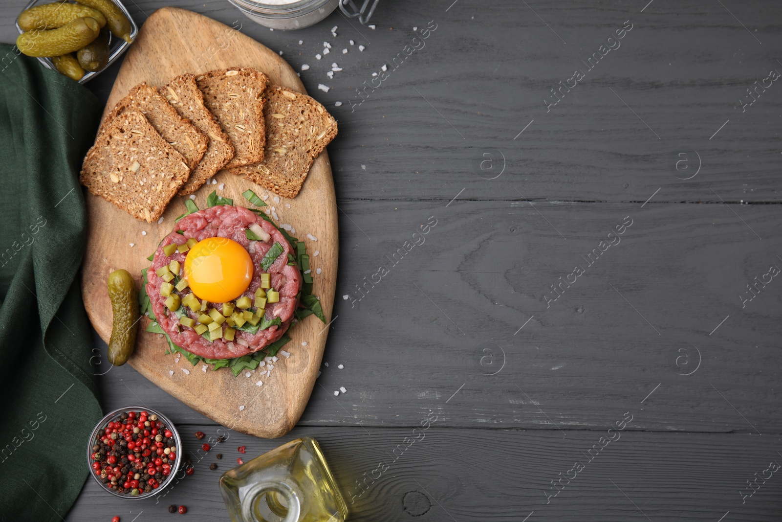 Photo of Tasty beef steak tartare served with yolk, pickled cucumber and other accompaniments on grey wooden table, flat lay. Space for text