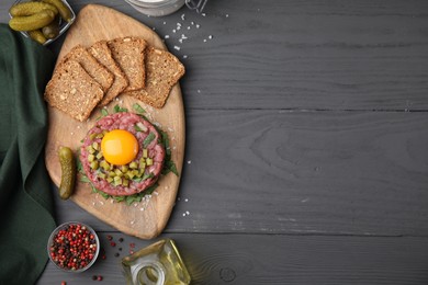 Photo of Tasty beef steak tartare served with yolk, pickled cucumber and other accompaniments on grey wooden table, flat lay. Space for text