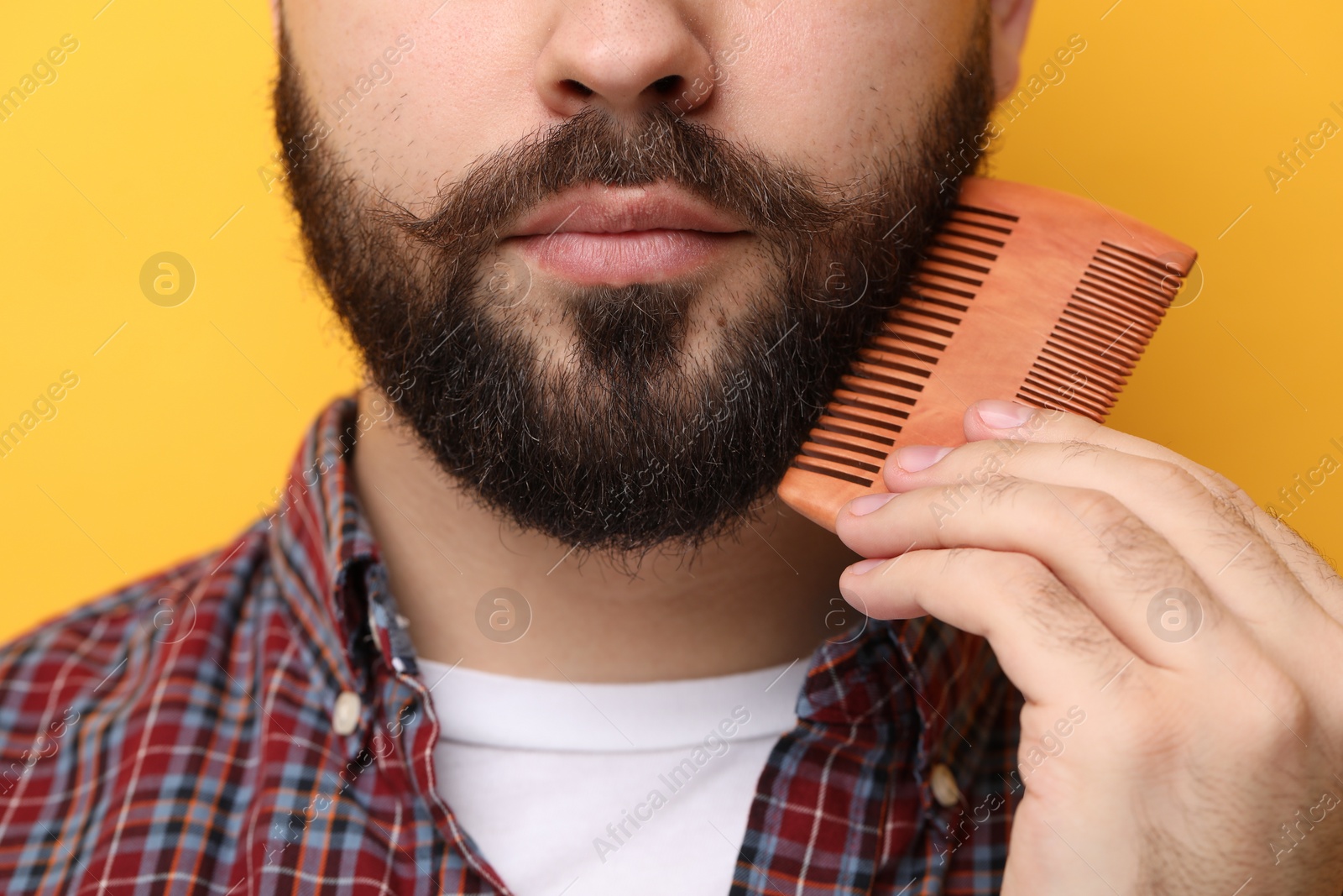 Photo of Handsome young man combing beard on yellow background, closeup