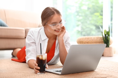 Young woman with glass of cola working on laptop at home. Refreshing drink