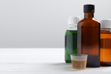 Bottles of syrup and measuring cup on white table against light grey background, space for text. Cold medicine