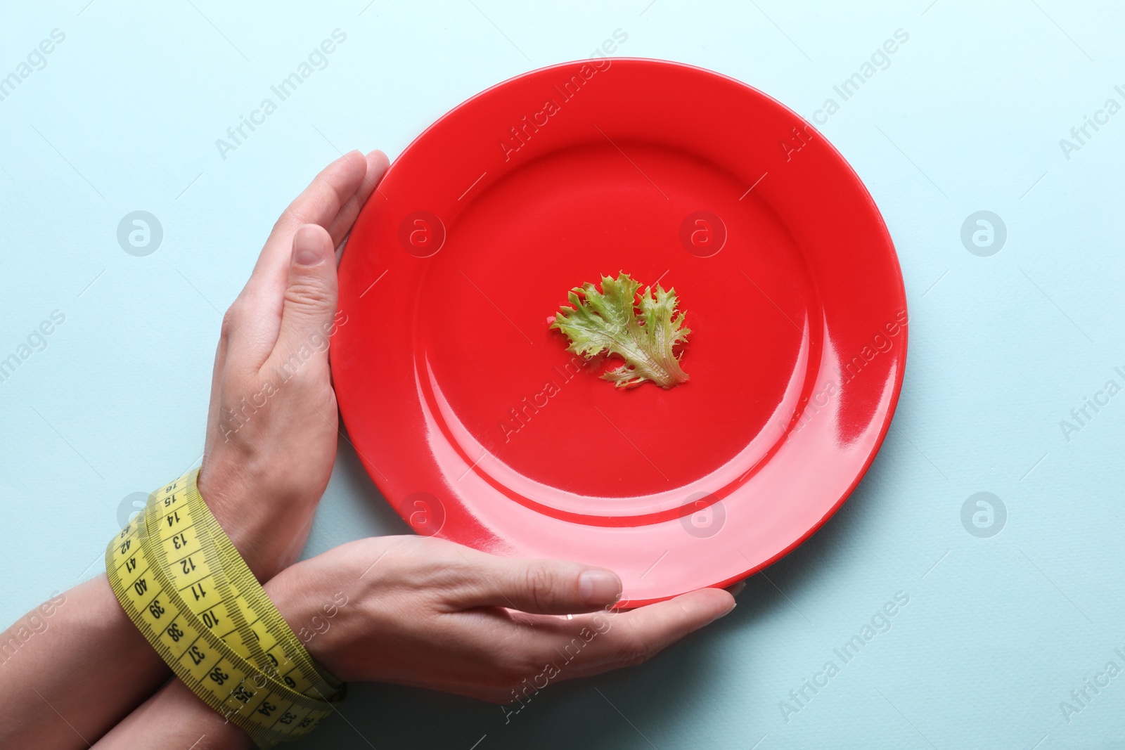 Photo of Woman holding plate with lettuce leaf in hands tied with measuring tape on light blue background, top view. Diet concept