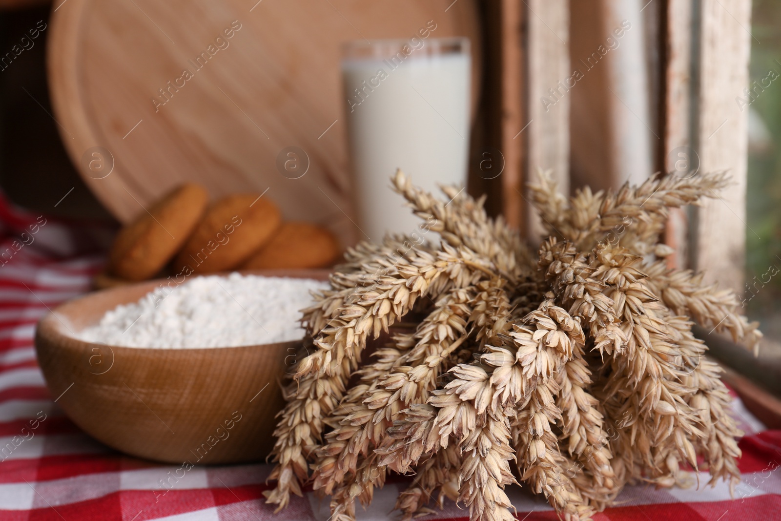 Photo of Bowl of wheat flour and spikes on red checkered tablecloth indoors