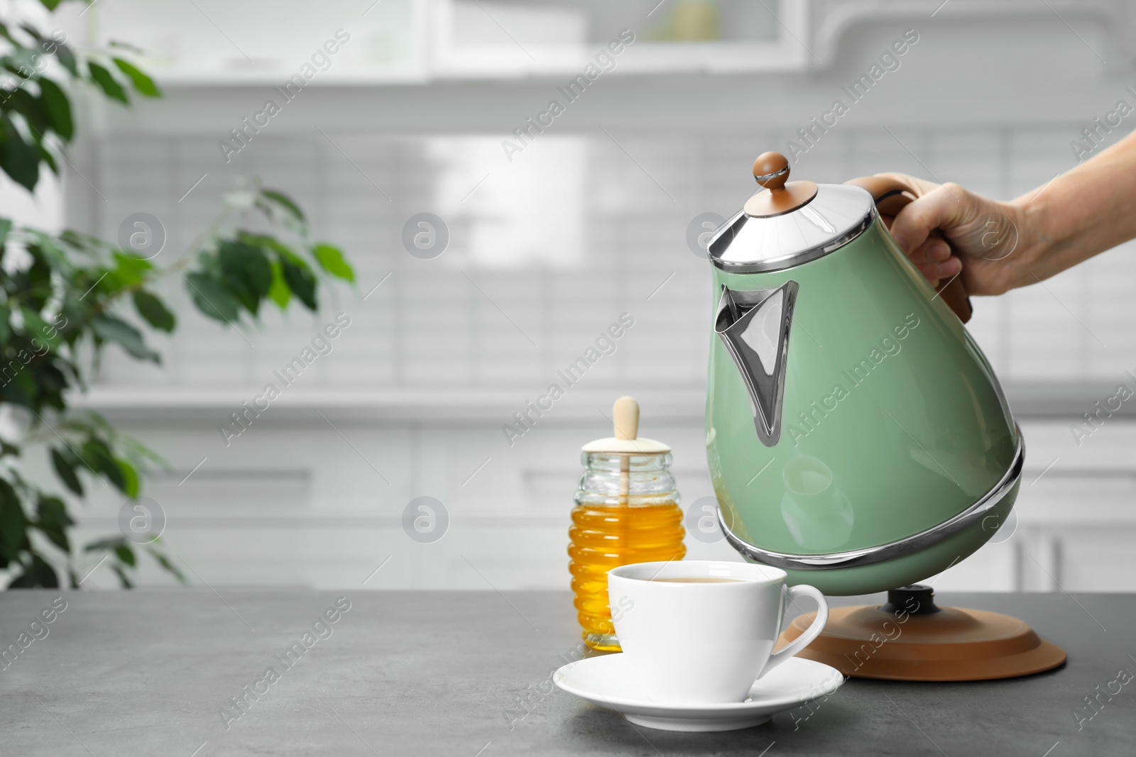 Photo of Woman pouring water from modern electric kettle into cup at grey table in kitchen, closeup. Space for text