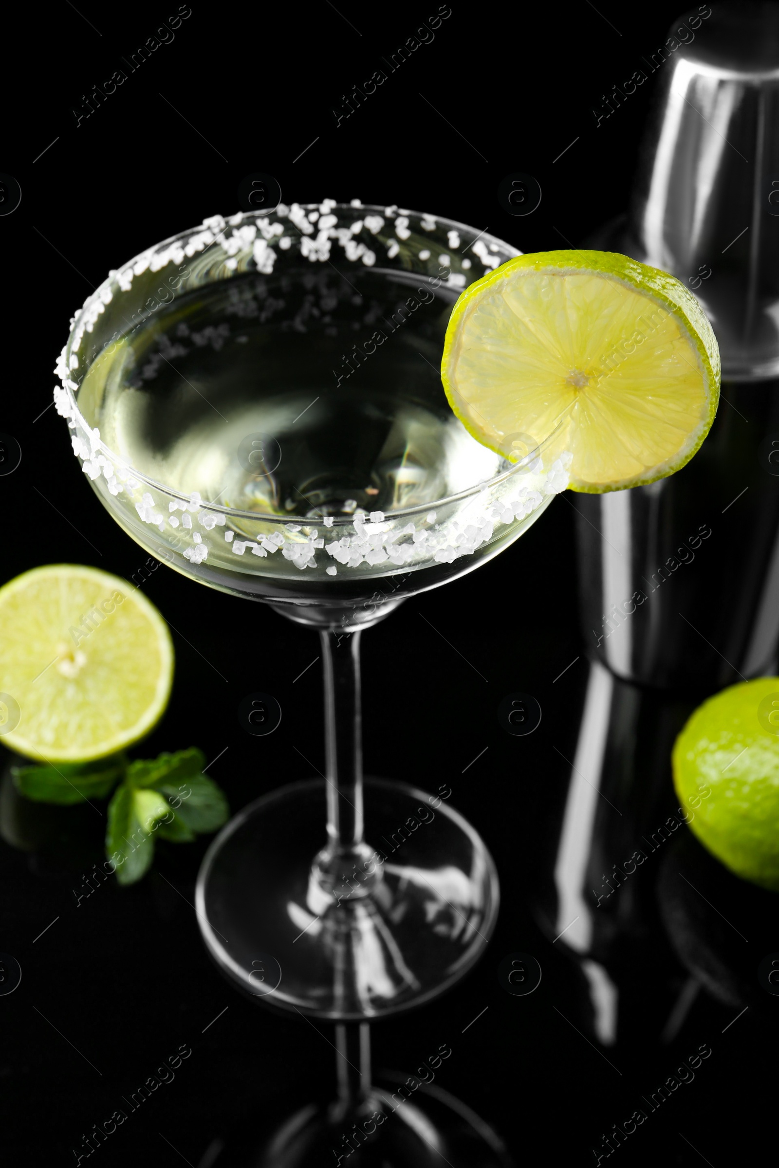 Photo of Delicious cocktail with salt, lime and metal shaker on black mirror surface