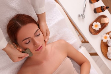Young woman receiving facial massage with jade gua sha tool in beauty salon, above view