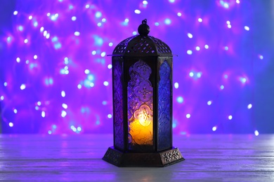 Photo of Muslim lamp with candle on table against blurred fairy lights. Fanous as Ramadan symbol