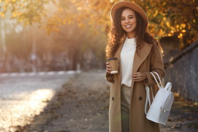 Photo of Portrait of beautiful African-American woman with stylish white backpack and hot drink on city street. Space for text