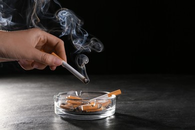 Photo of Woman holding smoldering cigarette over glass ashtray at grey table against black background, closeup