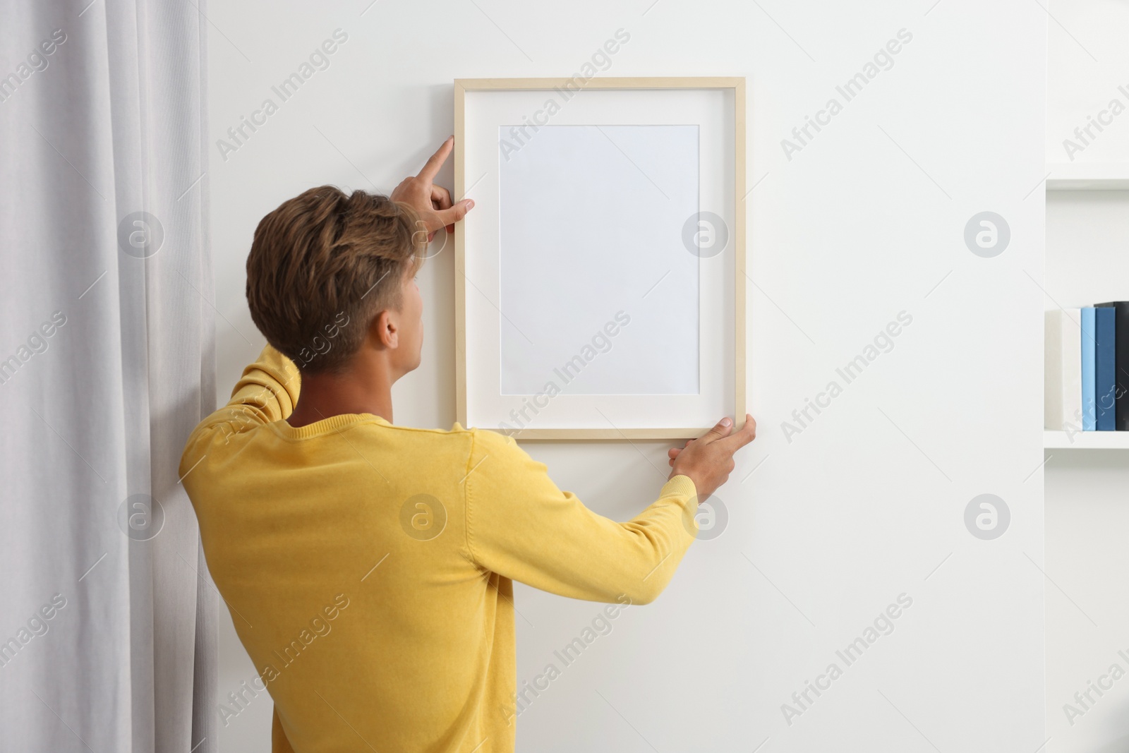 Photo of Young man hanging picture frame on white wall indoors, back view