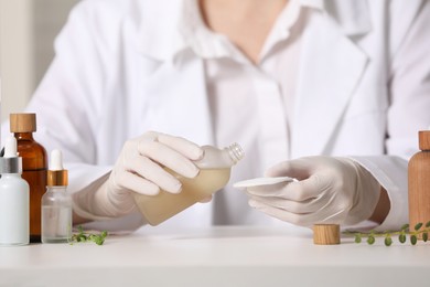 Dermatologist with bottle testing cosmetic product at white table indoors, selective focus