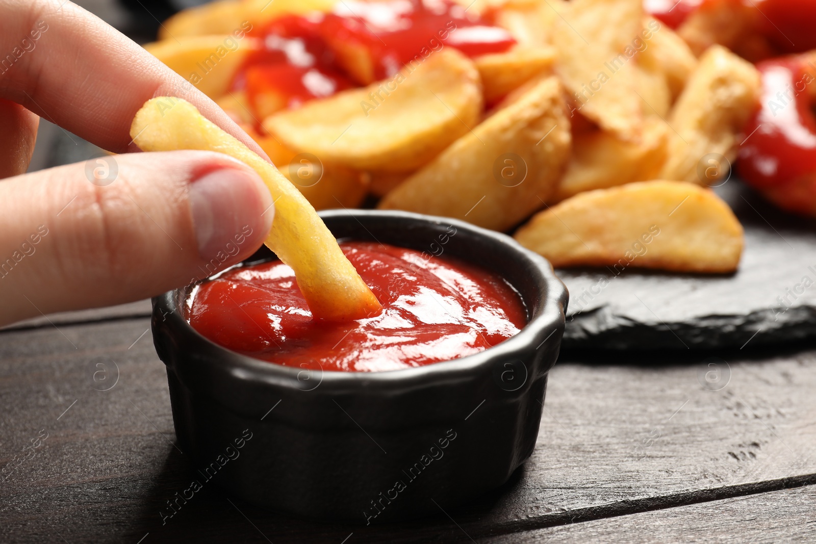 Photo of Woman dipping french fry into bowl with tasty ketchup at dark wooden table, closeup