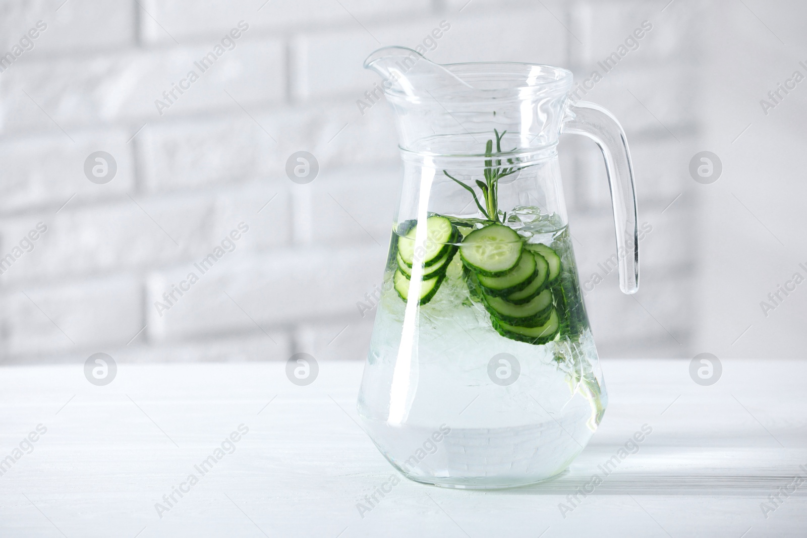 Photo of Refreshing cucumber water with rosemary in jug on white table against brick wall. Space for text