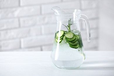 Refreshing cucumber water with rosemary in jug on white table against brick wall. Space for text