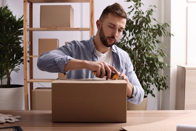 Man using utility knife to open parcel at wooden table indoors