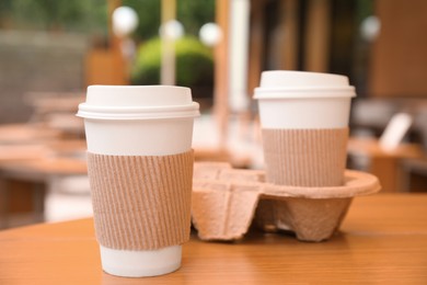 Photo of Takeaway paper coffee cups with plastic lids, sleeves and cardboard holder on wooden table outdoors