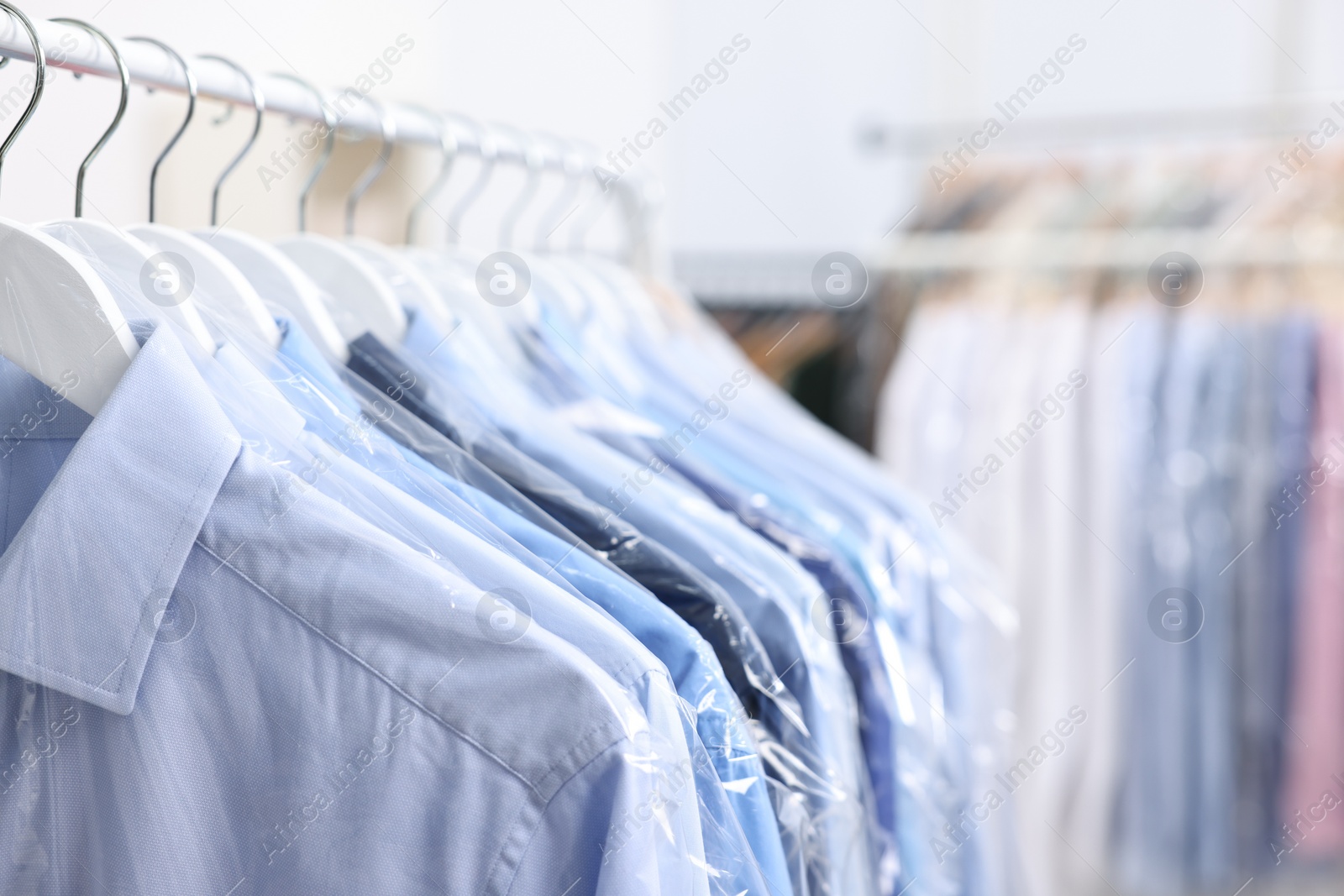 Photo of Dry-cleaning service. Many different clothes in plastic bags hanging on rack indoors, closeup