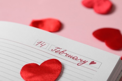Notebook with 14 February written on page corner against pink background, closeup. Valentine's Day celebration