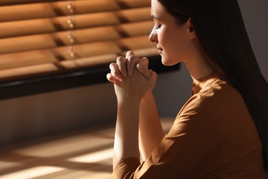 Religious young woman with clasped hands praying indoors