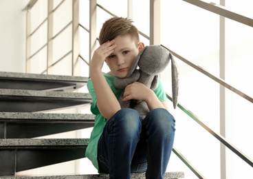 Sad little boy with toy sitting on stairs indoors