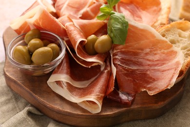 Photo of Slices of tasty cured ham, olives, bread and basil on wooden board, closeup