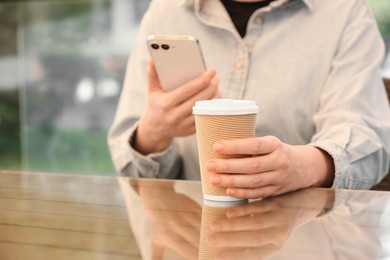 Photo of Woman holding takeaway paper cup and smartphone at table, closeup. Coffee to go