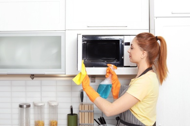 Woman cleaning microwave oven with rag and detergent in kitchen