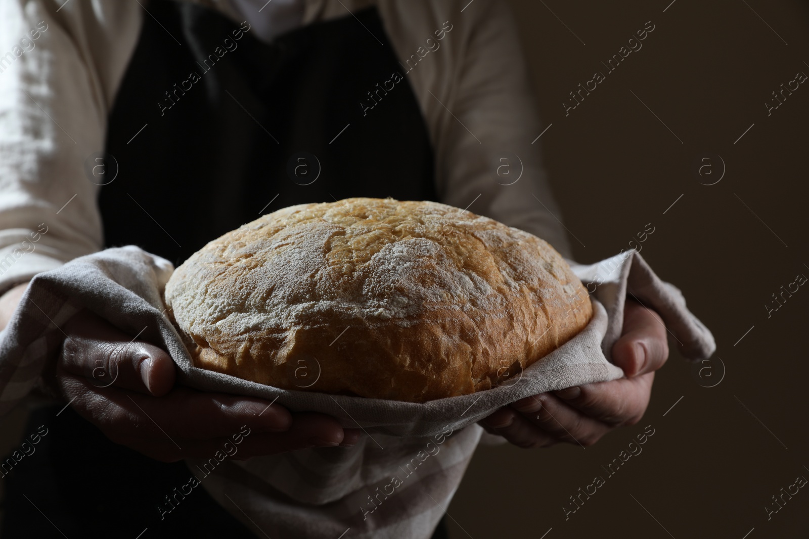 Photo of Man holding loaf of fresh bread on dark brown background, closeup