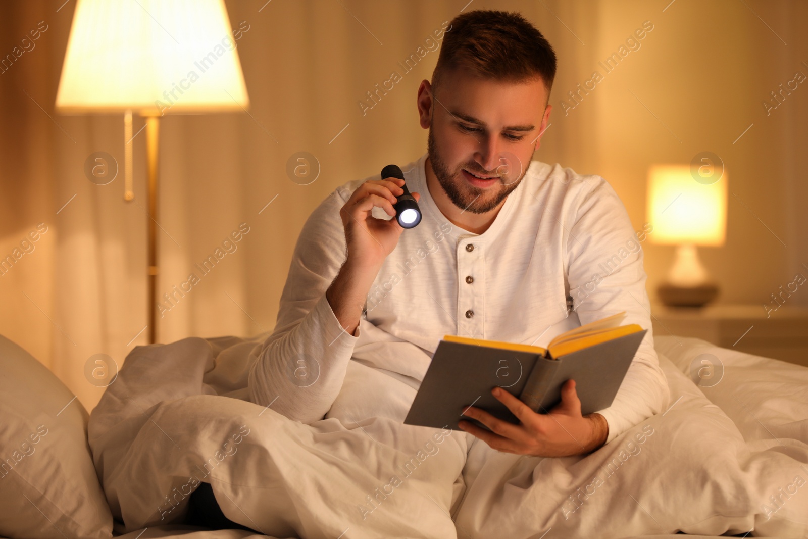 Photo of Young man with flashlight reading book in bedroom