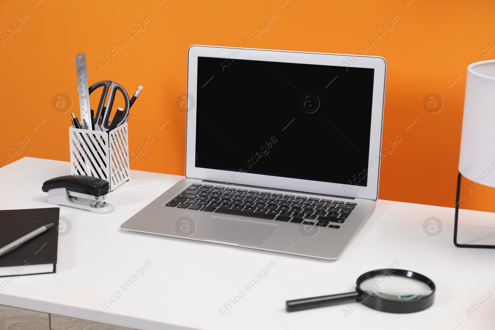 Photo of Workplace with laptop, stationery and magnifying glass on desk near orange wall. Home office