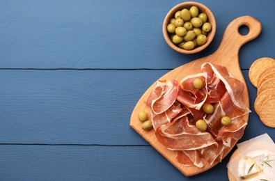 Photo of Slices of tasty cured ham, olives and cheese on blue wooden table, flat lay. Space for text